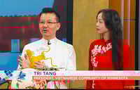 A man wearing Vietnamese traditional dress (áo dài) gestures with his hands and stands beside a woman also wearing áo dài. A screen a the wall of a TV studio are behind them.